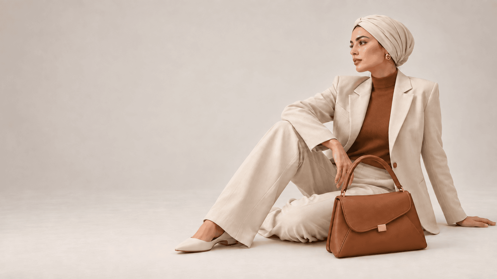 Model seated in a cream tailored suit and headwrap, holding a tan leather bag